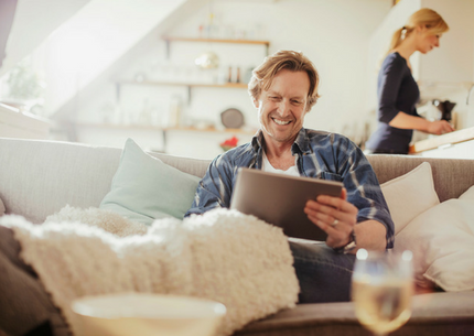 Man seating on the couch smiling at his tablet and a woman in the background in the kitchen