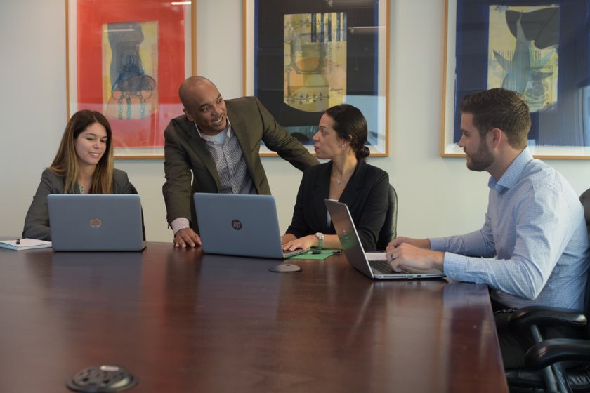 A group of people seating in a conference room around a table with their laptops