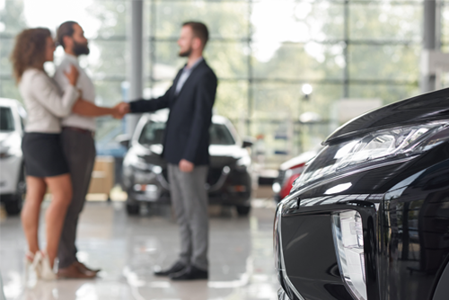 Car headlight, couple making deal with car dealer behind.
